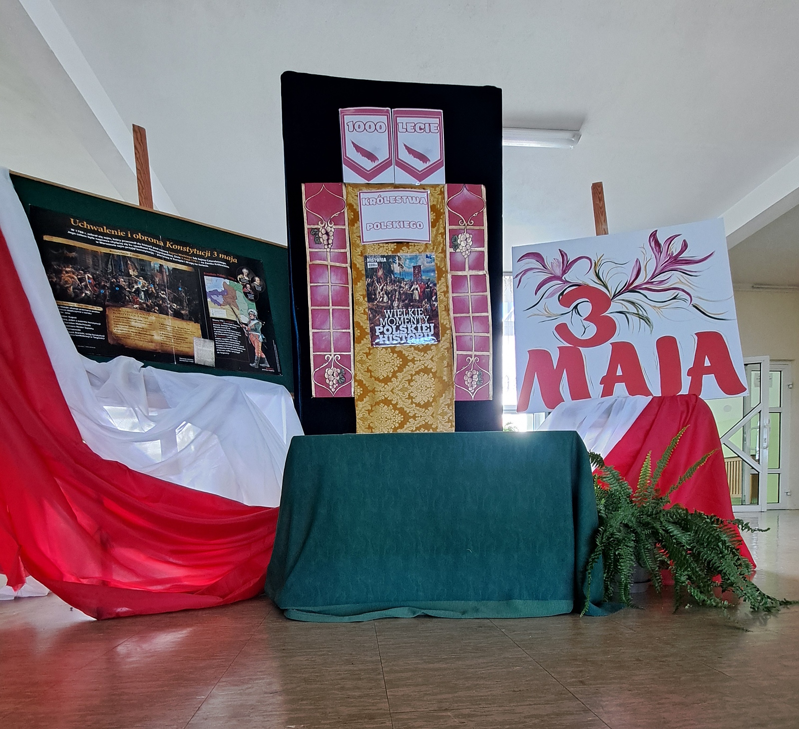 Dekoracja patriotyczna przygotowana w budynku szkoły. Sceneria związana jest z obchodami Święta 3 Maja. Dominują kolory biały i czerwony. Po lewej stronie znajduje się tablica informacyjna z ilustracją przedstawiającą scenę uchwalenia konstytucji. W środku stoi ozdobna plansza z napisem „1000-lecie Królestwa Polskiego” oraz reprodukcją obrazu. Po prawej stronie widnieje duży napis „3 Maja” ozdobiony dekoracyjnymi wzorami. Całość jest przystrojona biało-czerwoną tkaniną oraz paprocią na dole.