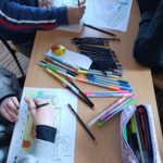 Widok pulpitu ławki. Na ławce kolorowanki, rozrzucone luźno kredki i flamastry. Otwarty piórnik. Widać dłonie uczniów podczas pracy.