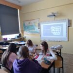 Cztery uczennice pochylone siedzące po dwóch stronach ławki. Praca na materiałami plastycznymi. W tle tablica multimedialna z logo programu.