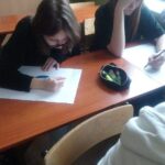 Dwie uczennica podczas pracy. Siedzą w ławce. Lekko pochylone nad białymi kartkami papieru.