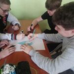 Trzech uczniów podczas przygotowywania jednej pracy rysunkowej. Siedzą po dwóch stronach ławki. W dłoniach kredki i markery. Na pierwszym planie otwarty piórnik.