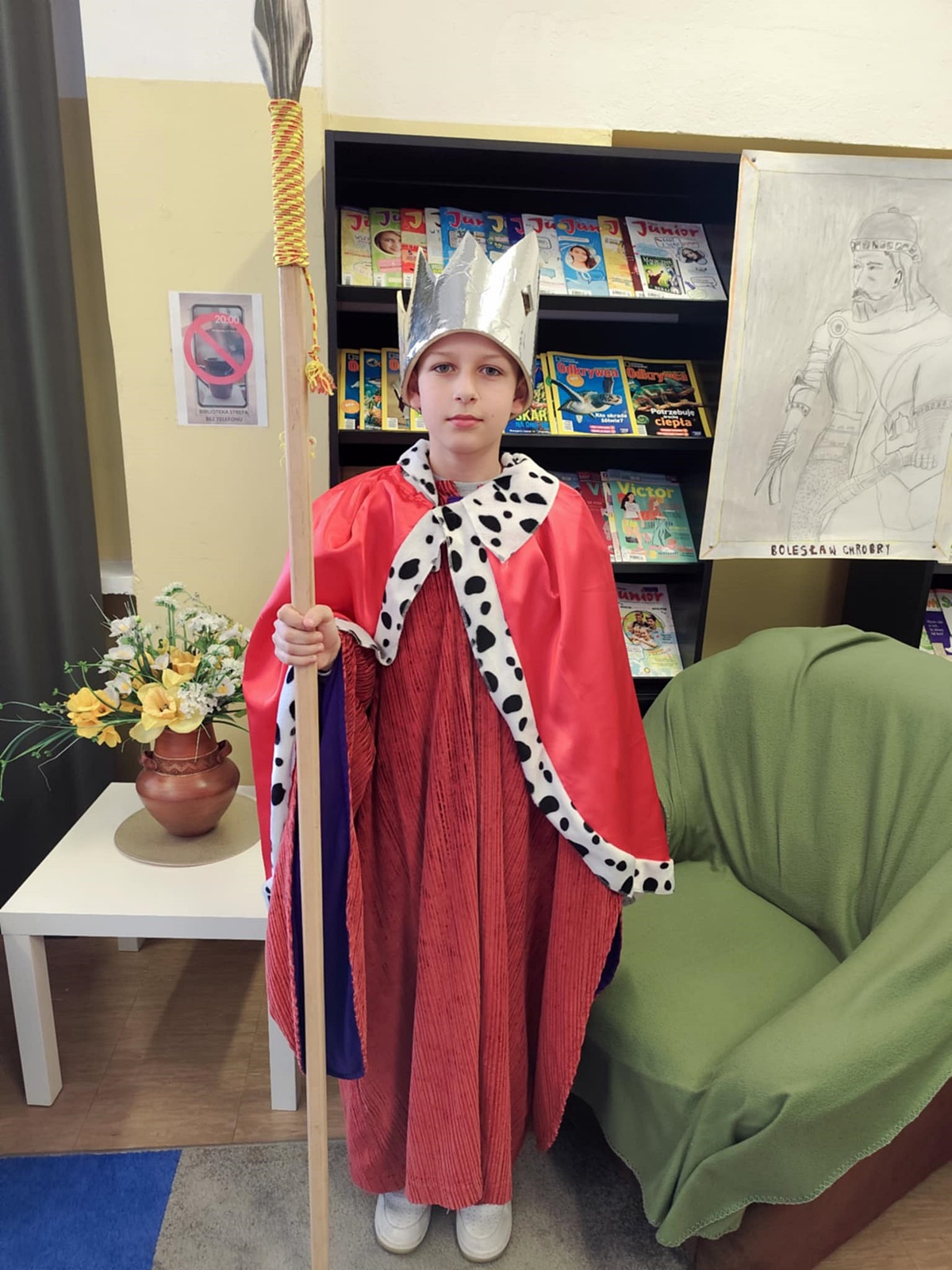 Uczeń przebrany za Bolesława Chrobrego. Czerwony strój. W prawej dłoni długa włócznia, na głowie duża srebrna korona. W tle stolik z kwiatami, zielony fotel i biblioteczne regały.