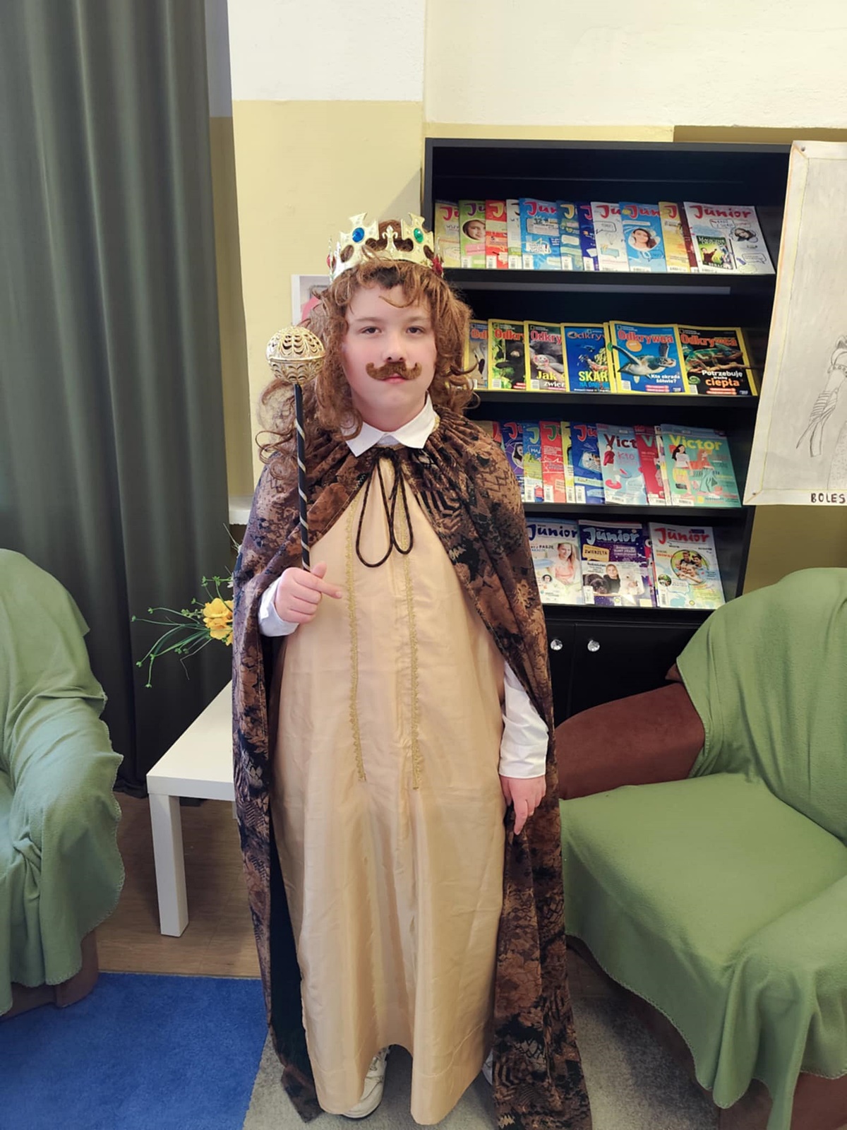 Uczeń przebrany za Bolesława Chrobrego. Brązowa peleryna, beżowy strój. Na głowie perka i korona. Na twarzy sztuczne wąsy. W tle wyposażenie biblioteki.