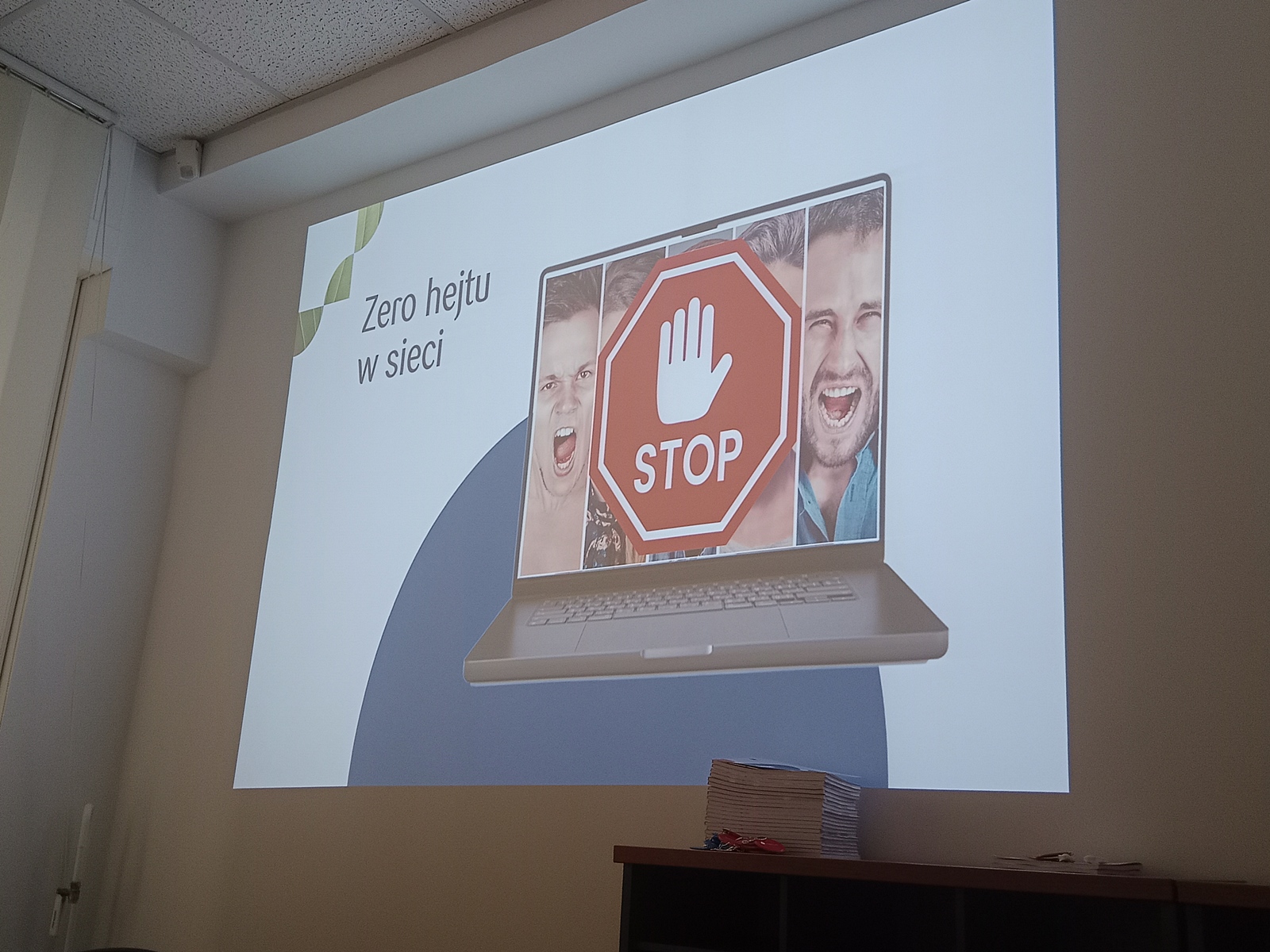Slajd z prezentacji wyświetlany na ścianie. Na slajdzie widać grafikę laptopa, na którego ekranie są trzy zdjęcia osób wyrażających złość lub agresję, a na środku znajduje się czerwony znak stopu z białą dłonią i napisem „STOP”. Po lewej stronie widnieje napis w języku polskim: „Zero hejtu w sieci”.