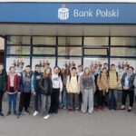 Grupa nastolatków stojących przed oddziałem banku PKO Bank Polski. Młodzież pozuje do zdjęcia razem z dwiema dorosłymi kobietami. Wszyscy stoją na chodniku przed budynkiem z dużym niebieskim szyldem „PKO Bank Polski”. Na szybach w tle widać plakaty z dziećmi trzymającymi książki i dyplomy.