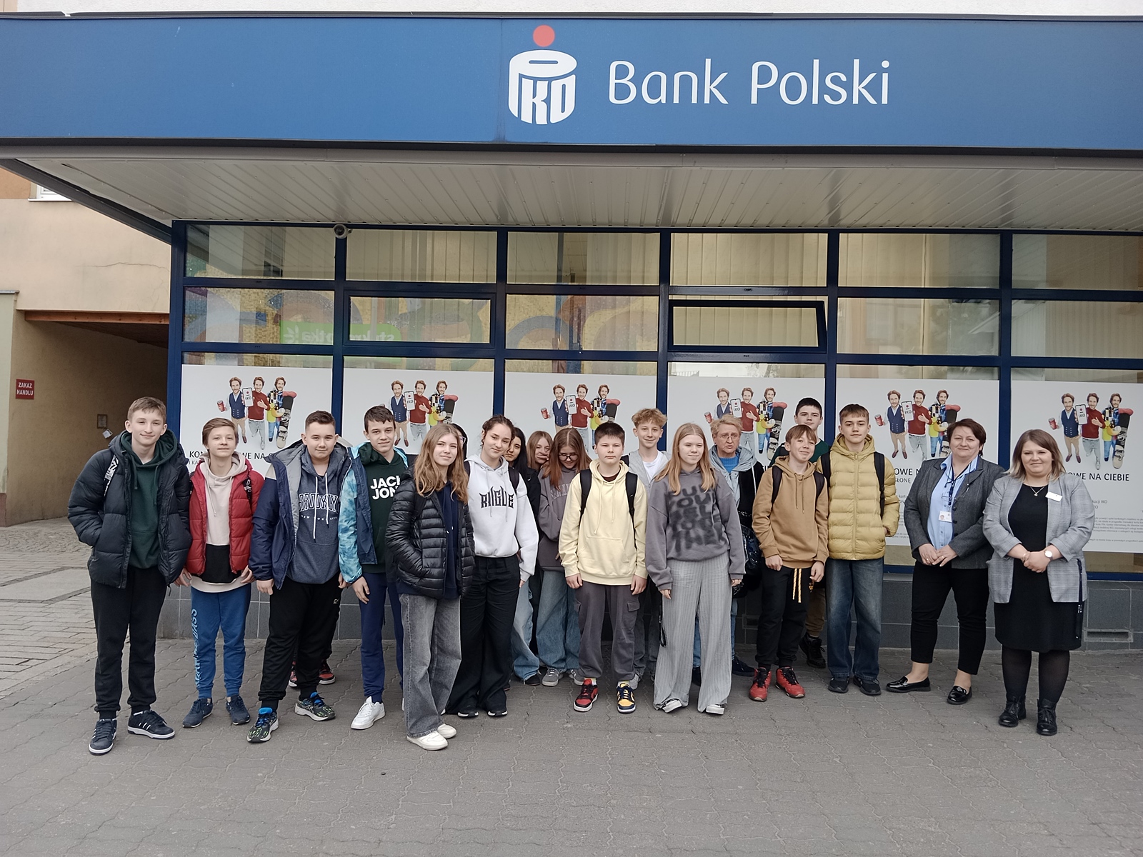 Grupa nastolatków stojących przed oddziałem banku PKO Bank Polski. Młodzież pozuje do zdjęcia razem z dwiema dorosłymi kobietami. Wszyscy stoją na chodniku przed budynkiem z dużym niebieskim szyldem „PKO Bank Polski”. Na szybach w tle widać plakaty z dziećmi trzymającymi książki i dyplomy.