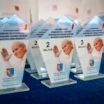 Szklane trofea ustawione w rzędach na granatowym obrusie. Każde trofeum przedstawia zdjęcie uśmiechniętego papieża Jana Pawła II z uniesioną ręką. Nagrody są opisane jako pamiątki z VI Powiatowego Konkursu Recytatorskiego poświęconego pamięci papieża. Na górze trofeum znajduje się cytat Jana Pawła II: „Wy jesteście przyszłością świata… Wy jesteście moją nadzieją!”.