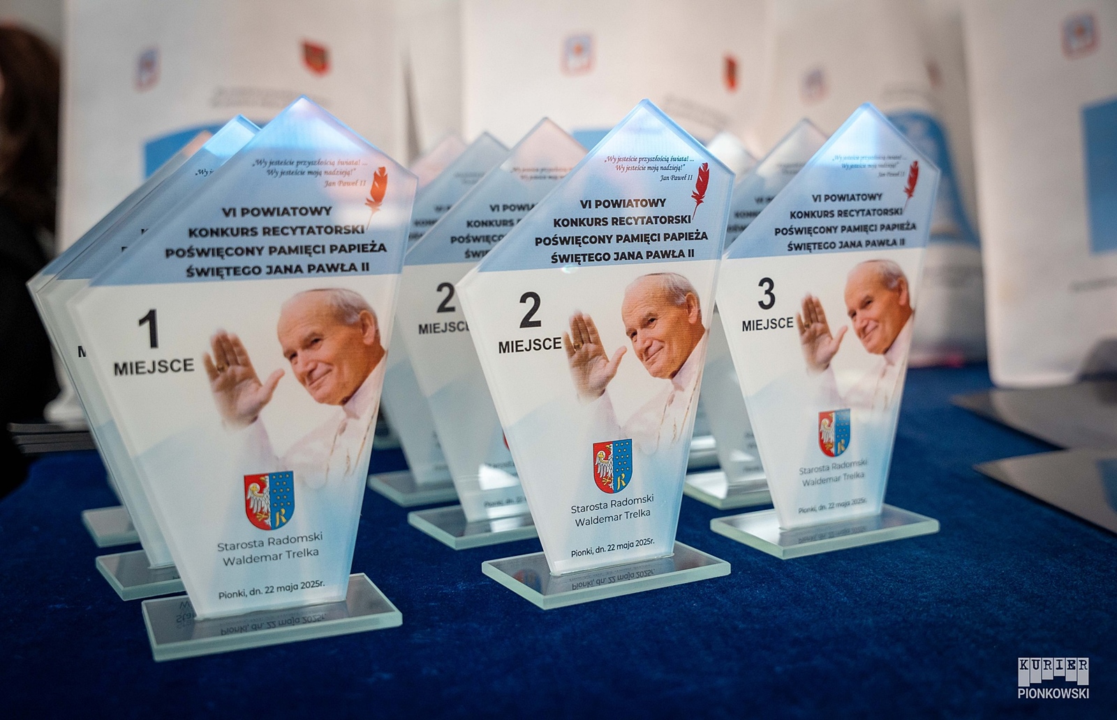 Szklane trofea ustawione w rzędach na granatowym obrusie. Każde trofeum przedstawia zdjęcie uśmiechniętego papieża Jana Pawła II z uniesioną ręką. Nagrody są opisane jako pamiątki z VI Powiatowego Konkursu Recytatorskiego poświęconego pamięci papieża. Na górze trofeum znajduje się cytat Jana Pawła II: „Wy jesteście przyszłością świata… Wy jesteście moją nadzieją!”.