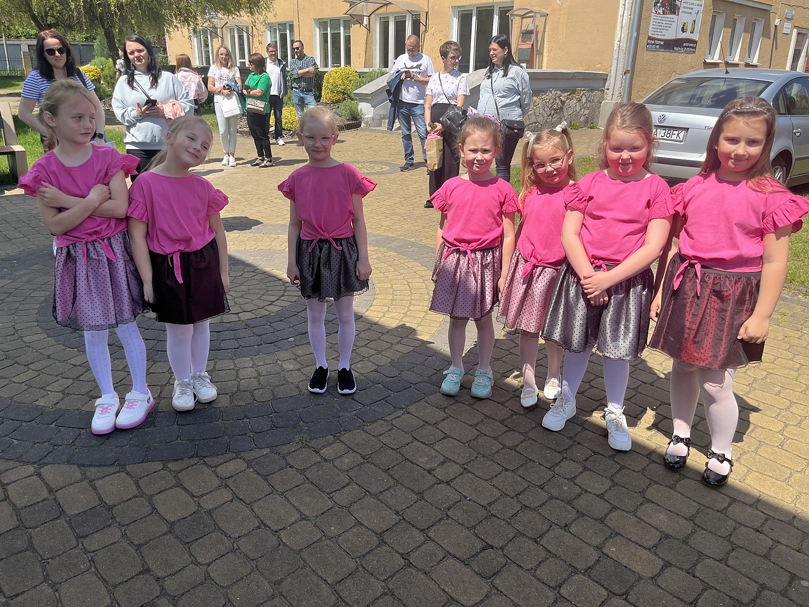Siedem małych dziewczynek, 1 klasa Szkoła Podstawowa, w różowych T-shirtach i różowych spodniczkach stoi przed występem na scenie. W tle rodzice dzieci.