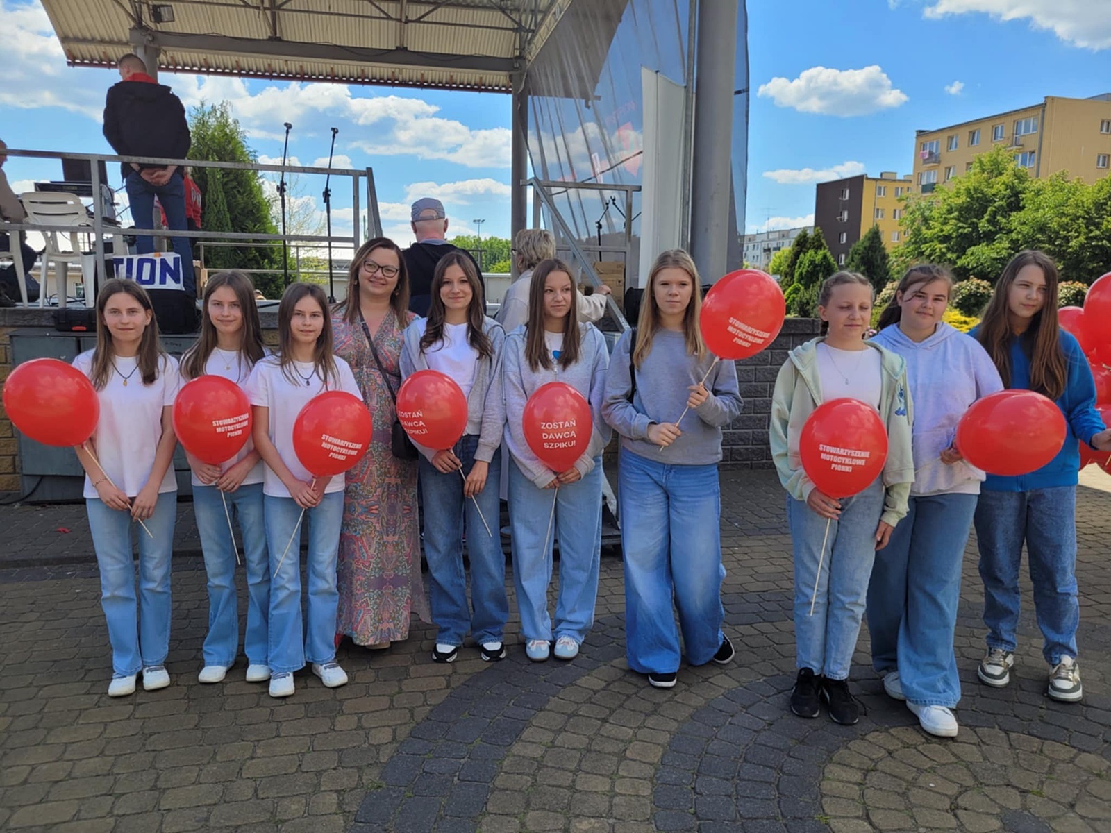 Dziewięć dziewczynek na tle sceny, ubranych w białe T-shirty i dżinsy , w ręce czerwone balony. W środku stoi ch opiekunka, w długiej kolorowej sukni, ma włosy do ramion, okulary.