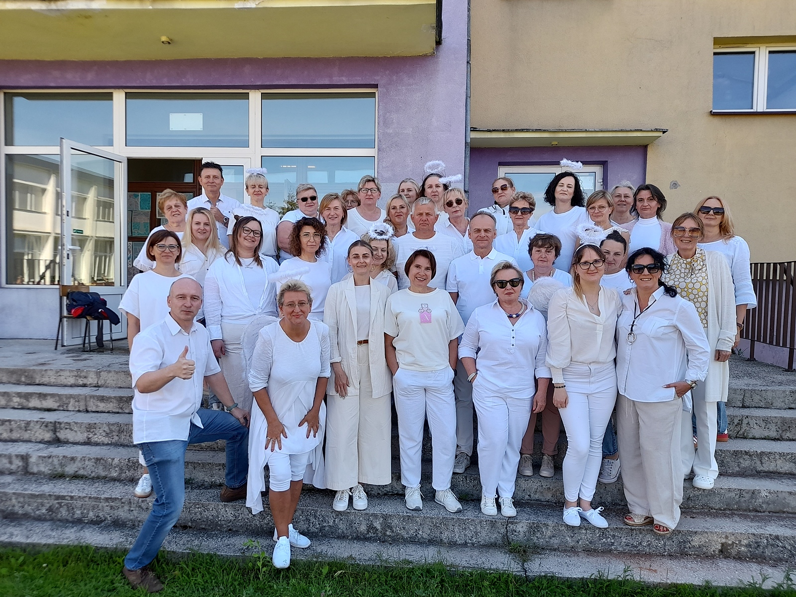 Duża grupa nauczycieli, stojących na schodach przed budynkiem szkoły. Wszyscy są ubrani na biało — część osób ma też opaski z anielskimi aureolami na głowie (biel symbolizuje „anielską cierpliwość” nauczycieli). Osoby pozują do wspólnego zdjęcia, uśmiechają się i wyglądają na zadowolone. W tle widać fioletowo-żółtą ścianę budynku z dużymi oknami i otwartymi drzwiami wejściowymi. Panuje pogodna, słoneczna atmosfera.