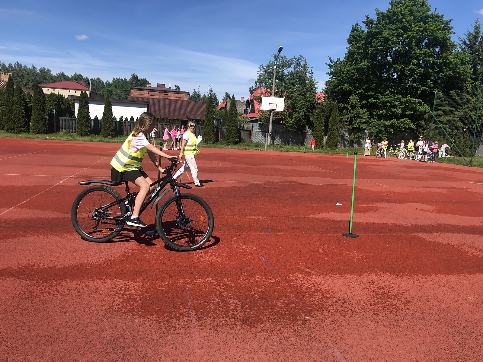 Dziewczynka jadącą na rowerze po czerwonym boisku sportowym. Ma na sobie różową koszulkę i kamizelkę odblaskową. Obok niej stoi kobieta również w kamizelce, nadzorująca przebieg praktycznego egzaminu na Kartę rowerową. W tle, przy ogrodzeniu, stoi grupa dzieci z rowerami, czekających na swoją kolej. Pogoda jest słoneczna, niebo bezchmurne, a wokół boiska rośnie dużo zielonych drzew.