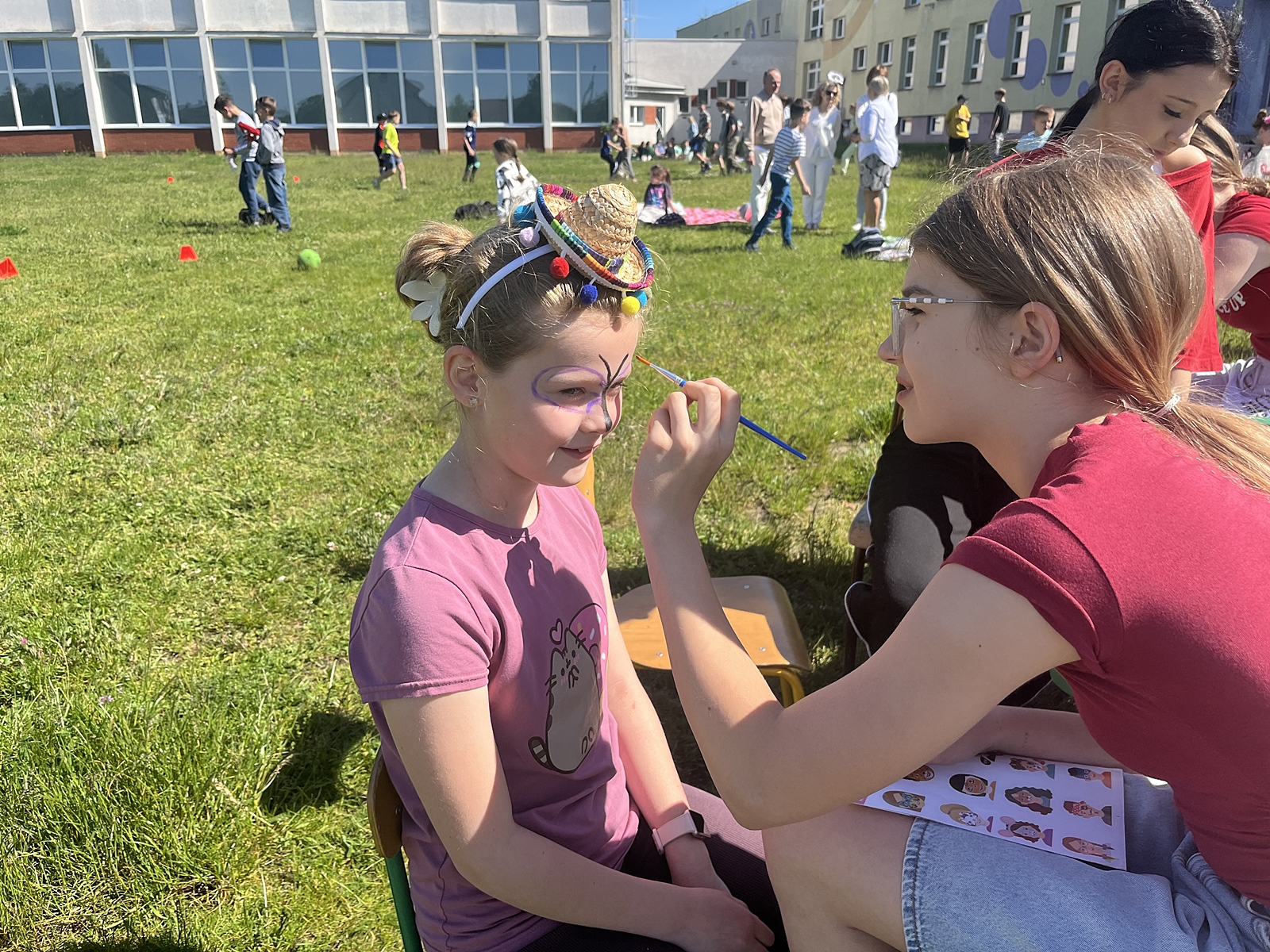 Starsza dziewczynka wykonuje rysunek na twarzy młodszej (malowanie twarzy). W tle budynek szkoły oraz zabawy sportowe na trawie grupy dzieci. Panuje pogodna i słoneczna atmosfera.