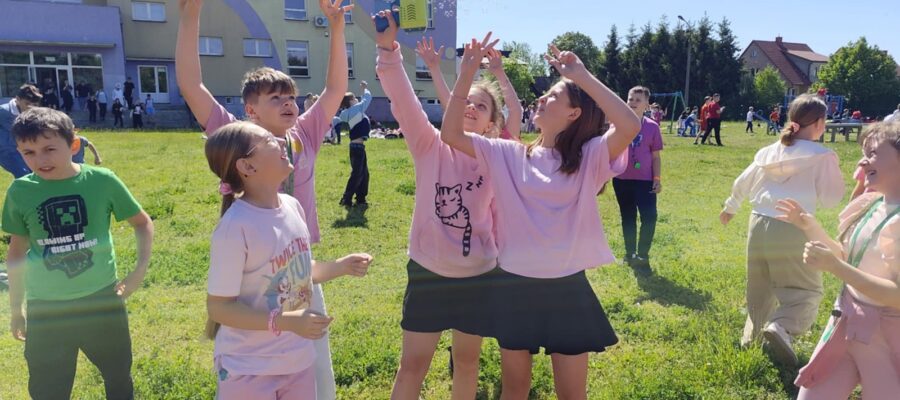 Grupa dzieci bawiących się na zielonej trawie przed budynkiem szkoły. Kilkoro dzieci podnosi ręce w górę, łapiąc bańki mydlane z automatu trzymanego przez jedną z dziewczynek. Panuje radosna atmosfera, dzieci się uśmiechają, a pogoda jest słoneczna i ciepła. Większość dzieci ma na sobie kolorowe ubrania, zwłaszcza różowe i zielone. W tle widać inne dzieci oraz dorosłych.