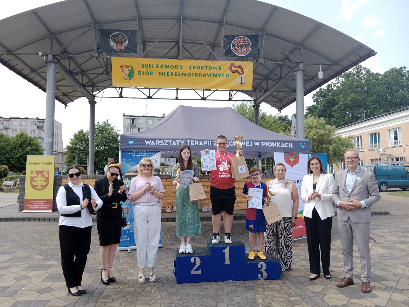 Podium z trojgiem dzieci— dwoma chłopcami i dziewczynką — które zajęły odpowiednio pierwsze, drugie i trzecie miejsce w zawodach sportowych dla osób z niepełnosprawnościami. Dzieci trzymają dyplomy i puchary. Za nimi stoi grupa dorosłych. W tle widać duży baner z napisem: „XVIII Zawody Sportowe Osób Niepełnosprawnych” oraz namiot z napisem „Warsztaty Terapii Zajęciowej w Pionkach”. Zdjęcie zostało zrobione na zewnątrz, pod zadaszoną sceną.