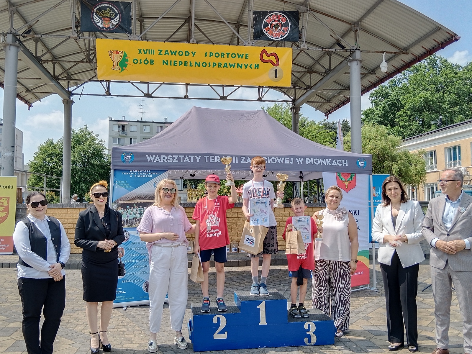 Podium z trzema chłopcami – uczestnikami XVIII Zawodów Sportowych Osób Niepełnosprawnych. Stoją na miejscach pierwszym, drugim i trzecim, trzymając puchary, dyplomy i torby z nagrodami. Za nimi znajduje się namiot z napisem „Warsztaty Terapii Zajęciowej w Pionkach” oraz baner zawodów. Po bokach podium stoją dorośli.