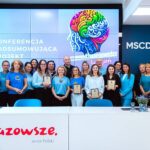 Grupa 20 osób stojących razem w jasnym, nowoczesnym pomieszczeniu konferencyjnym. Wiele z nich trzyma w rękach tabliczki z wyróżnieniami. W tle widoczny jest duży ekran z kolorową grafiką mózgu oraz napisami w języku polskim: „Konferencja podsumowująca projekt” i „Tydzień różnorodności”. Po prawej stronie widać logo MSCDN (Mazowieckie Samorządowe Centrum Doskonalenia Nauczycieli). Panuje radosna i uroczysta atmosfera.