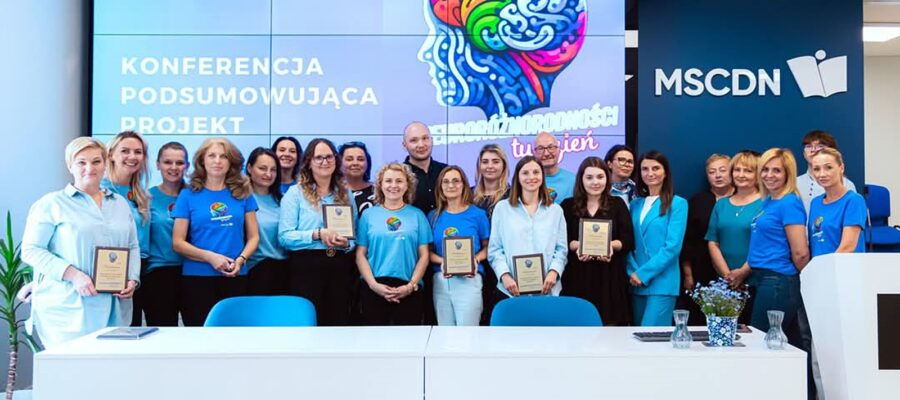 Grupa 20 osób stojących razem w jasnym, nowoczesnym pomieszczeniu konferencyjnym. Wiele z nich trzyma w rękach tabliczki z wyróżnieniami. W tle widoczny jest duży ekran z kolorową grafiką mózgu oraz napisami w języku polskim: „Konferencja podsumowująca projekt” i „Tydzień różnorodności”. Po prawej stronie widać logo MSCDN (Mazowieckie Samorządowe Centrum Doskonalenia Nauczycieli). Panuje radosna i uroczysta atmosfera.