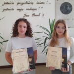Dwie dziewczynki stojące obok siebie na szkolnym korytarzu. Każda z nich trzyma w rękach dyplom oraz książkę jako nagrodę. Obie mają jasne bluzki. W tle widać tablicę pamiątkową z wizerunkiem Jana Pawła II oraz cytat na ścianie w języku polskim: „Wy jesteście przyszłością świata!... Wy jesteście moją nadzieją!” – Jan Paweł II.