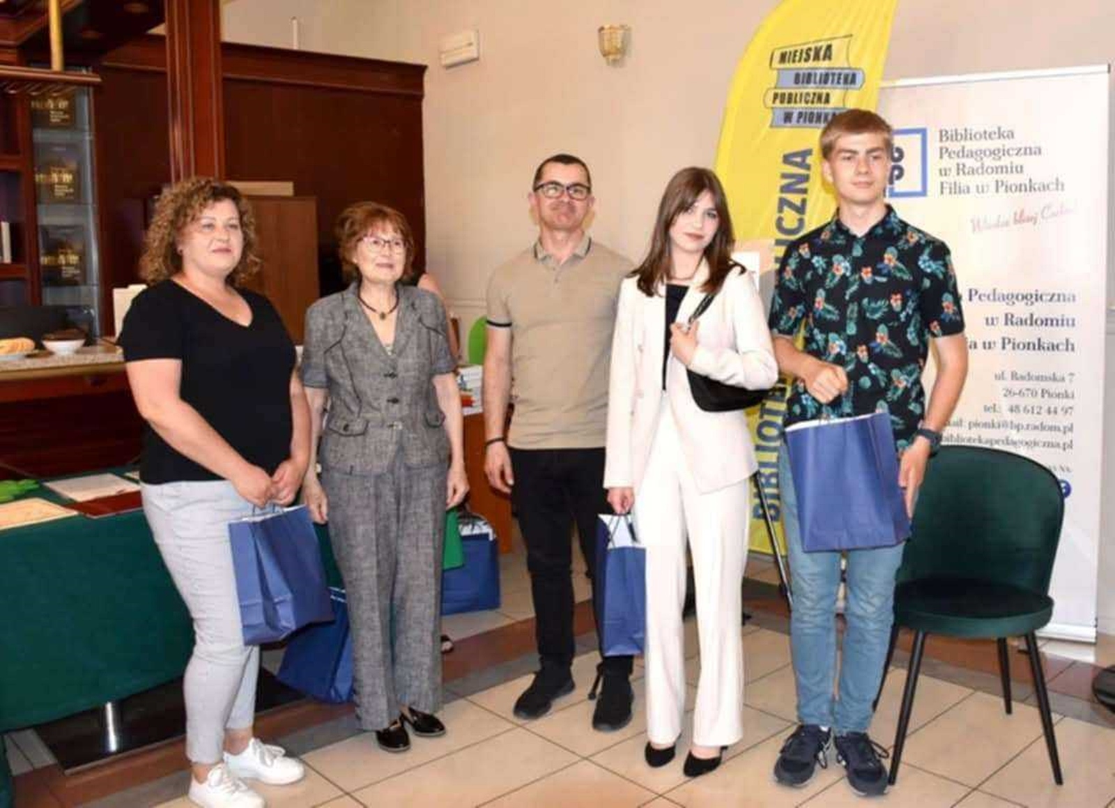 Pięć osób pozuje do wspólnego zdjęcia – organizatorzy konkursu i osoby odbierające nagrody. W tle widoczne są banery z nazwami bibliotek: „Miejska Biblioteka Publiczna w Pionkach” oraz „Biblioteka Pedagogiczna w Radomiu, Filia w Pionkach”. Atmosfera jest uroczysta i oficjalna.