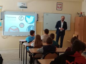 Mężczyzna prowadzi prelekcję w sali szkolnej z prezentacją dotyczącą podszywania się. W prezentacji znajdują się loga mediów społecznościowych.