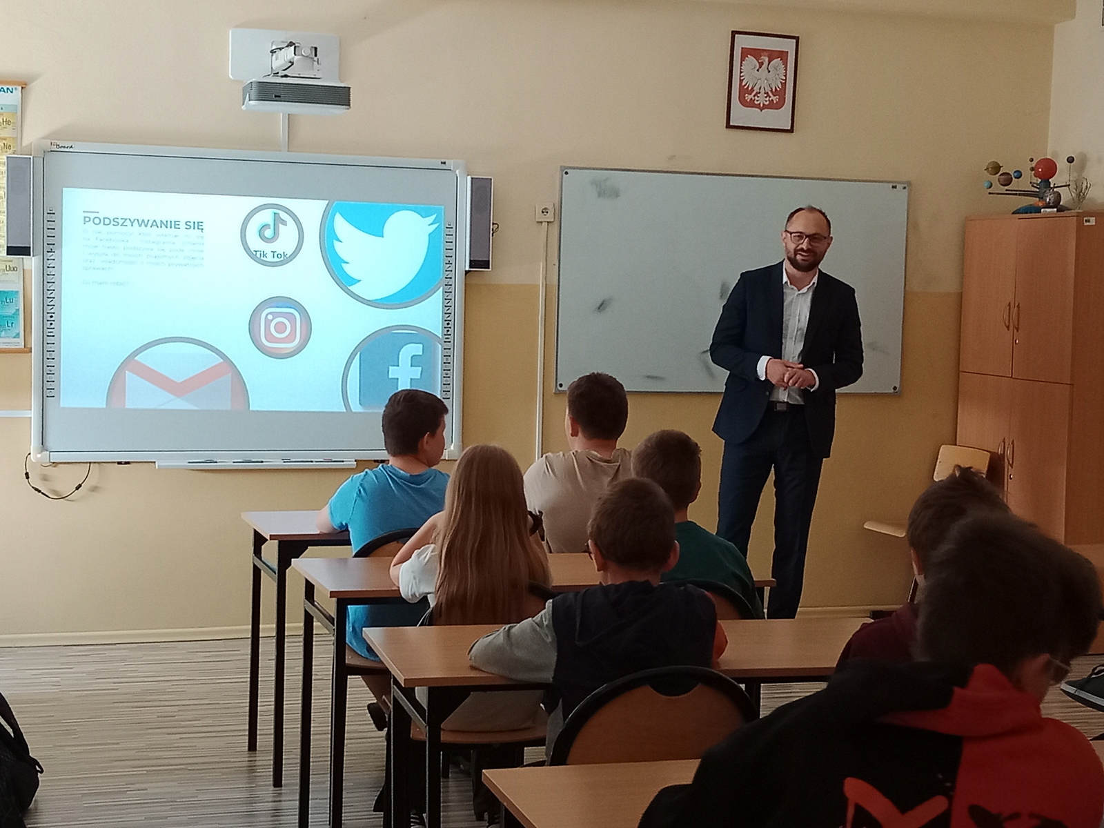 Mężczyzna prowadzi prelekcję w sali szkolnej z prezentacją dotyczącą podszywania się. W prezentacji znajdują się loga mediów społecznościowych.