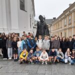 Zdjęcie grupowe. Uczniowie w dwóch rzędach. Rząd pierwszy w przysiadzie. W tle pomnik Jana Pawła II, fragment budynku kościoła i perspektywa kamienicy.