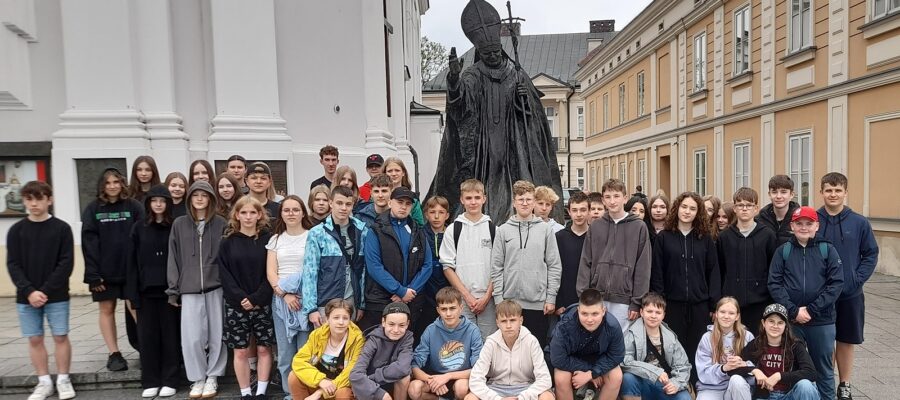 Zdjęcie grupowe. Uczniowie w dwóch rzędach. Rząd pierwszy w przysiadzie. W tle pomnik Jana Pawła II, fragment budynku kościoła i perspektywa kamienicy.