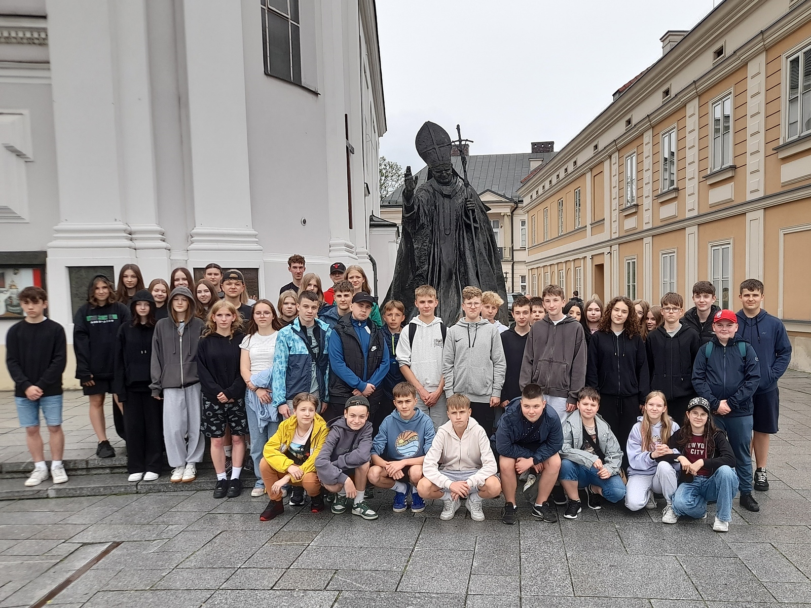 Zdjęcie grupowe. Uczniowie w dwóch rzędach. Rząd pierwszy w przysiadzie. W tle pomnik Jana Pawła II, fragment budynku kościoła i perspektywa kamienicy.