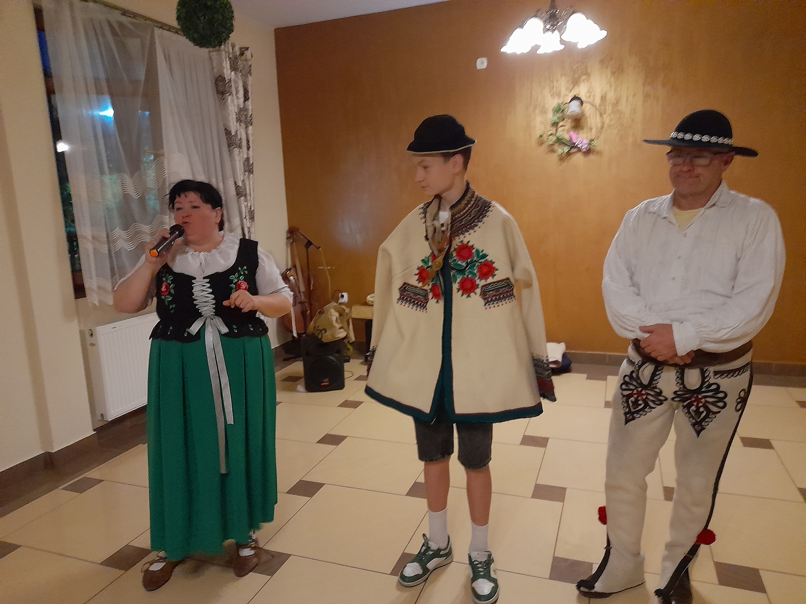 Kobieta i mężczyzna w strojach góralskich. Pomiędzy nimi uczeń w haftowanym płaszczu góralskim i kapeluszu. Kobieta trzyma w ręku mikrofon.