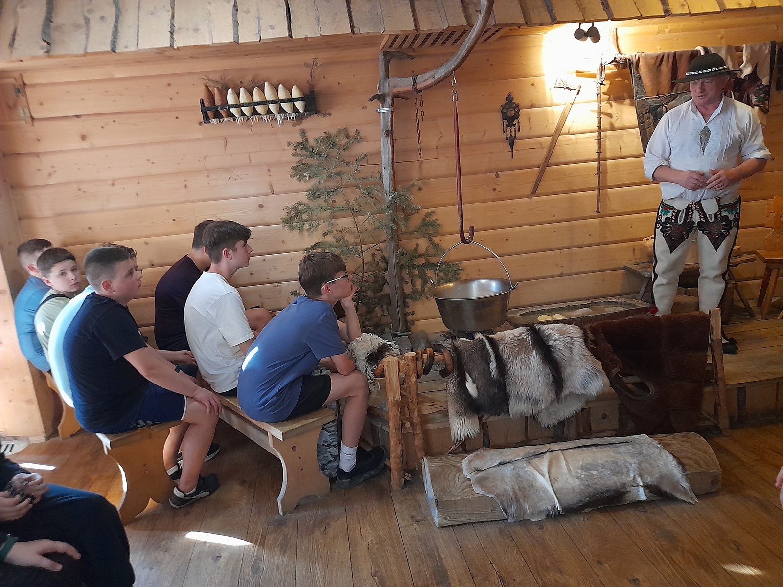 Wnętrze chaty góralskiej. Po lewej uczniowie na ławkach, z prawej góral opowiadający o wyrobie oscypków. Widoczne skóry zwierzęce, kociołek, oscypki pływające w jakimś płynie.