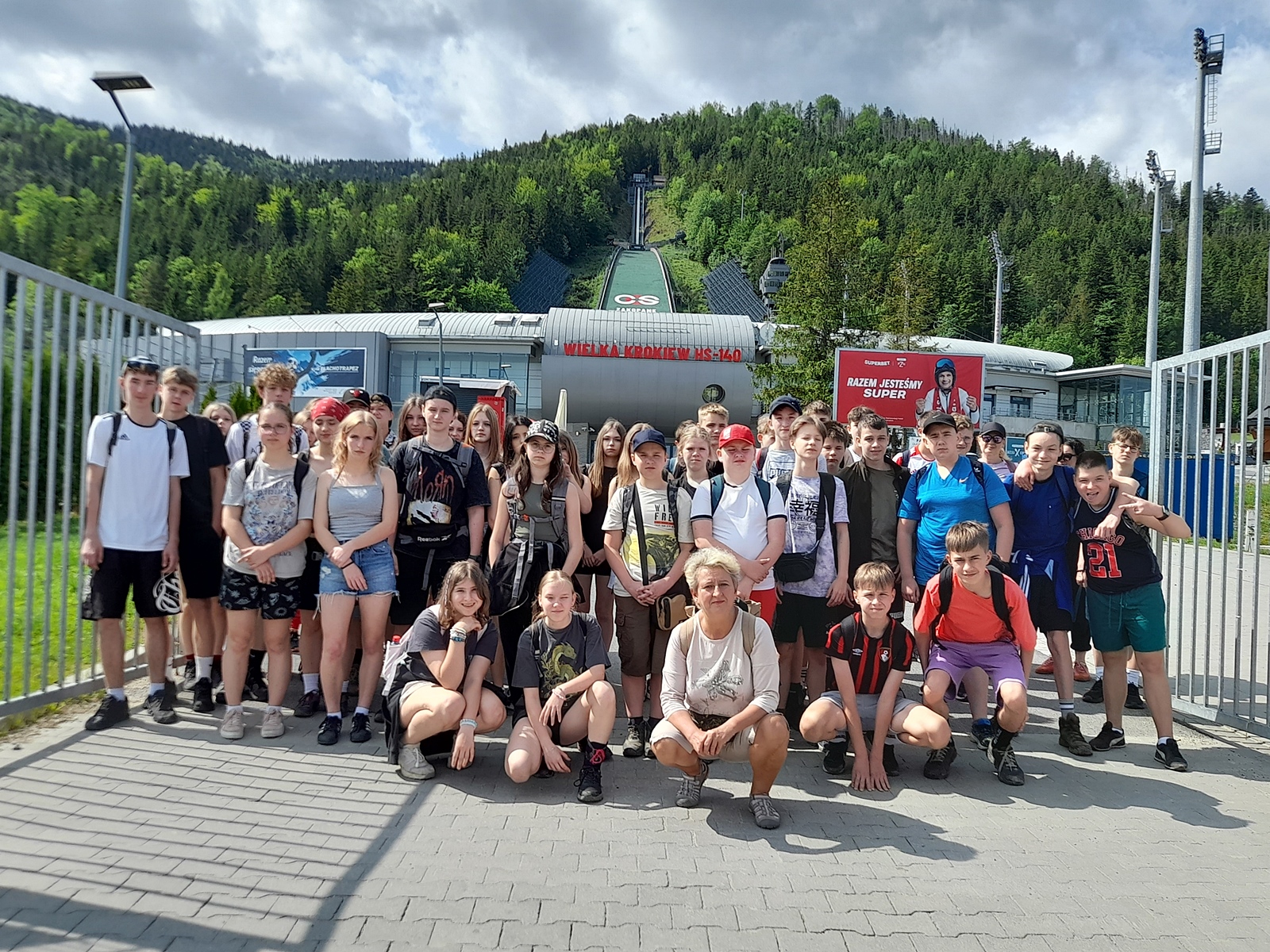 Zdjęcie grupowe pod skocznią. W tle skocznia i budynki z napisem "Wielka Krokiew HS-140".