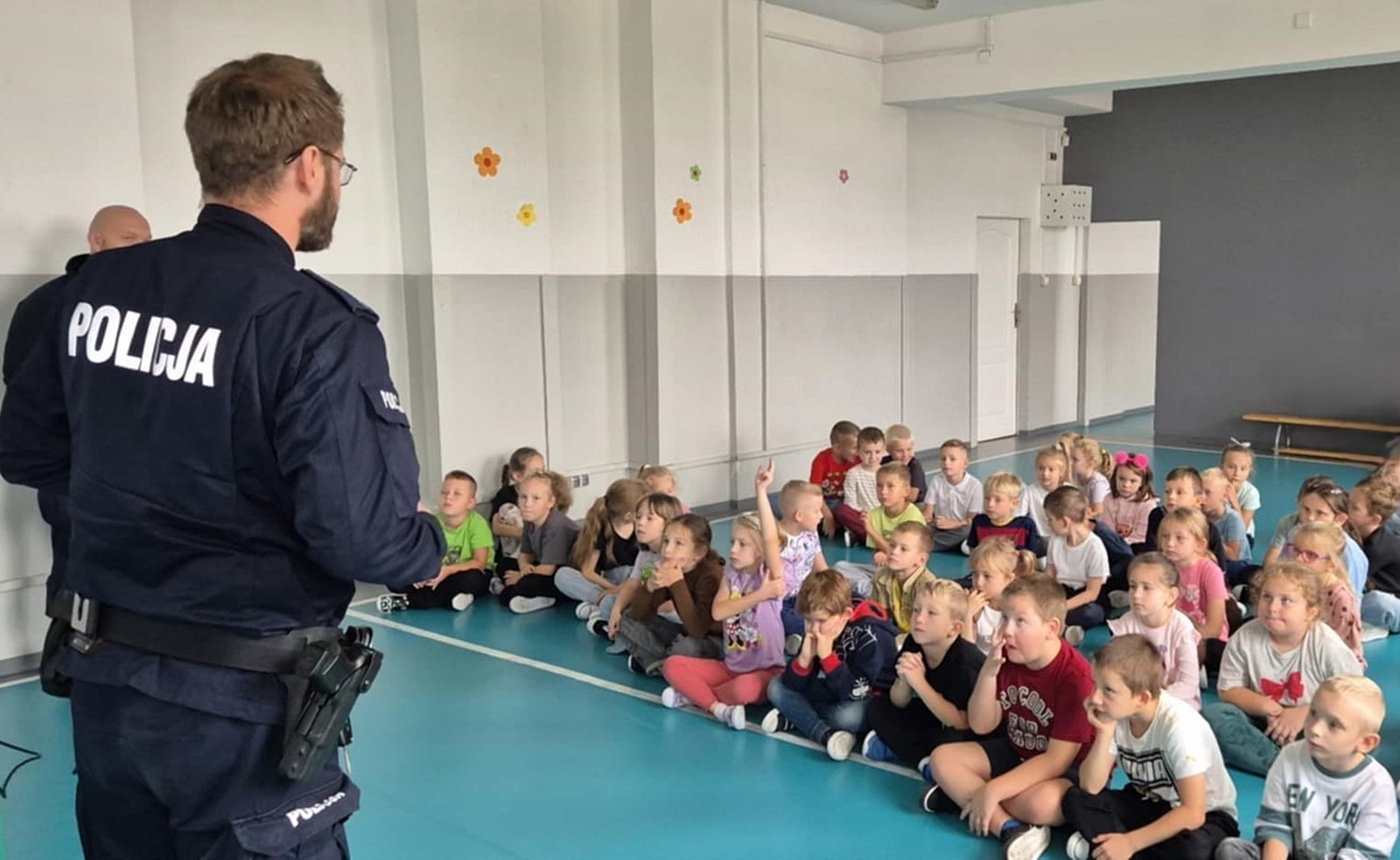 Grupa dzieci siedzących na podłodze w sali gimnastycznej i policjant w mundurze.