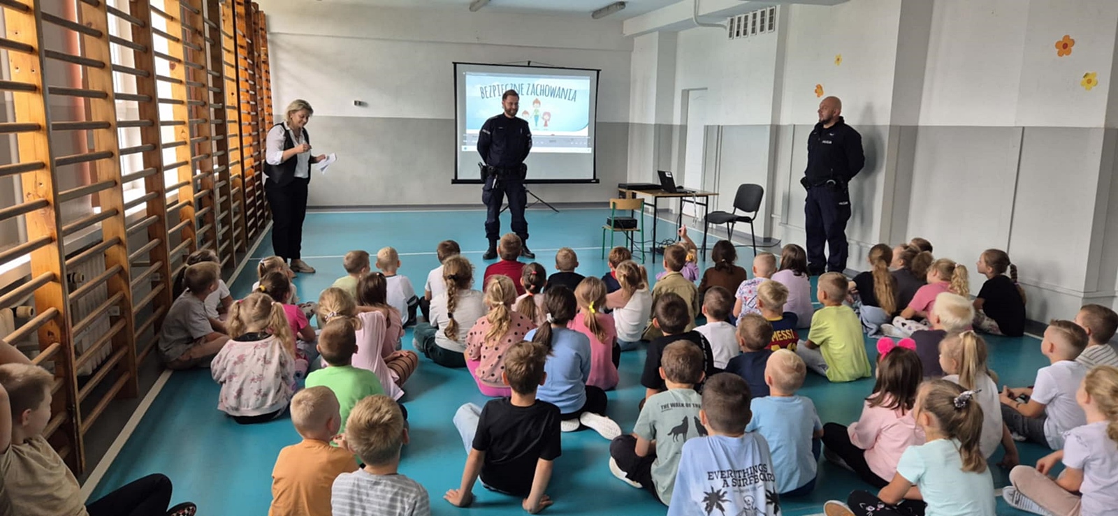 Grupa dzieci siedzących na podłodze w sali gimnastycznej, zwróconych twarzą do ekranu oraz dwóch policjantów w mundurach.