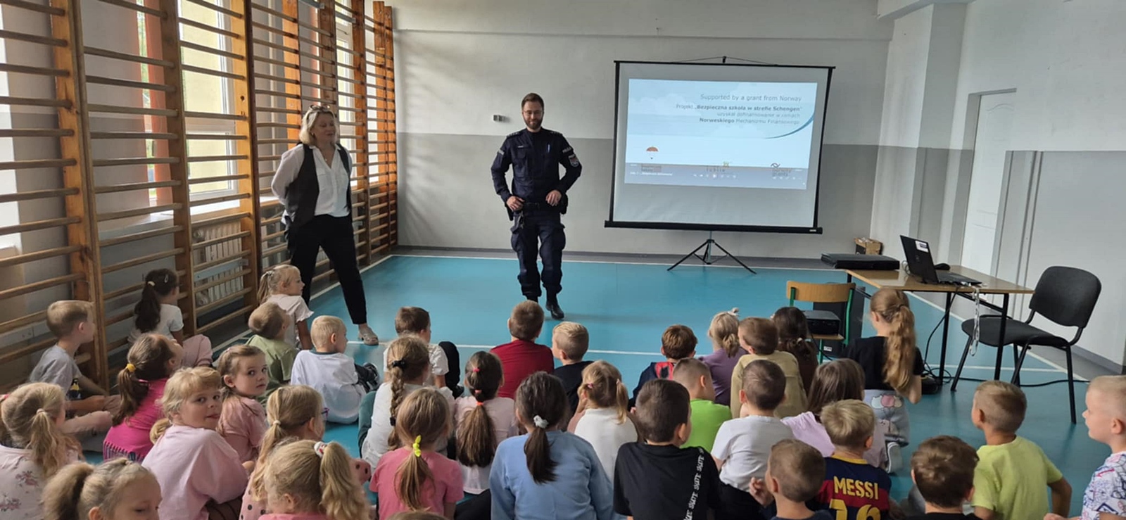 Grupa dzieci siedzących na podłodze w sali gimnastycznej, zwróconych twarzą do ekranu i dwóch dorosłych prowadzących spotkanie. Jeden z nich to policjant w mundurze. Za nimi znajduje się ekran z prezentacją, na której widnieje napis o projekcie „Bezpieczna szkoła w strefie Schengen”.