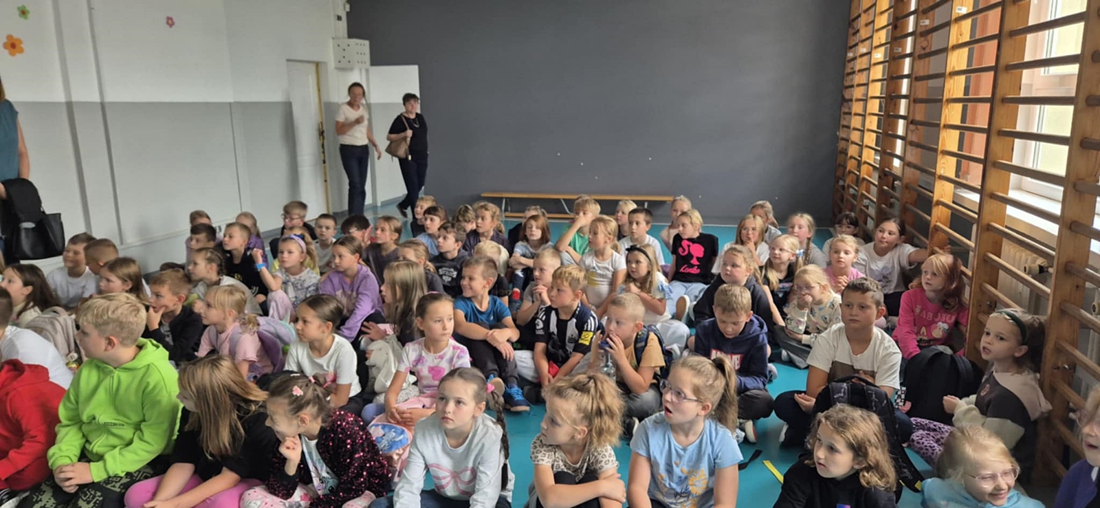 Duża grupa dzieci siedzących na podłodze w sali gimnastycznej.