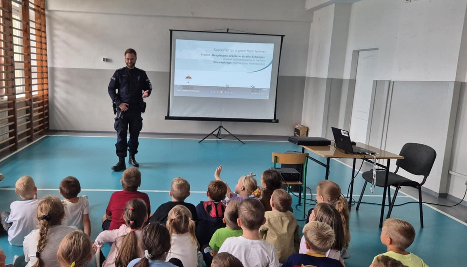 Grupa dzieci siedzących na podłodze w sali gimnastycznej, zwróconych twarzą do ekranu oraz policjant w mundurze