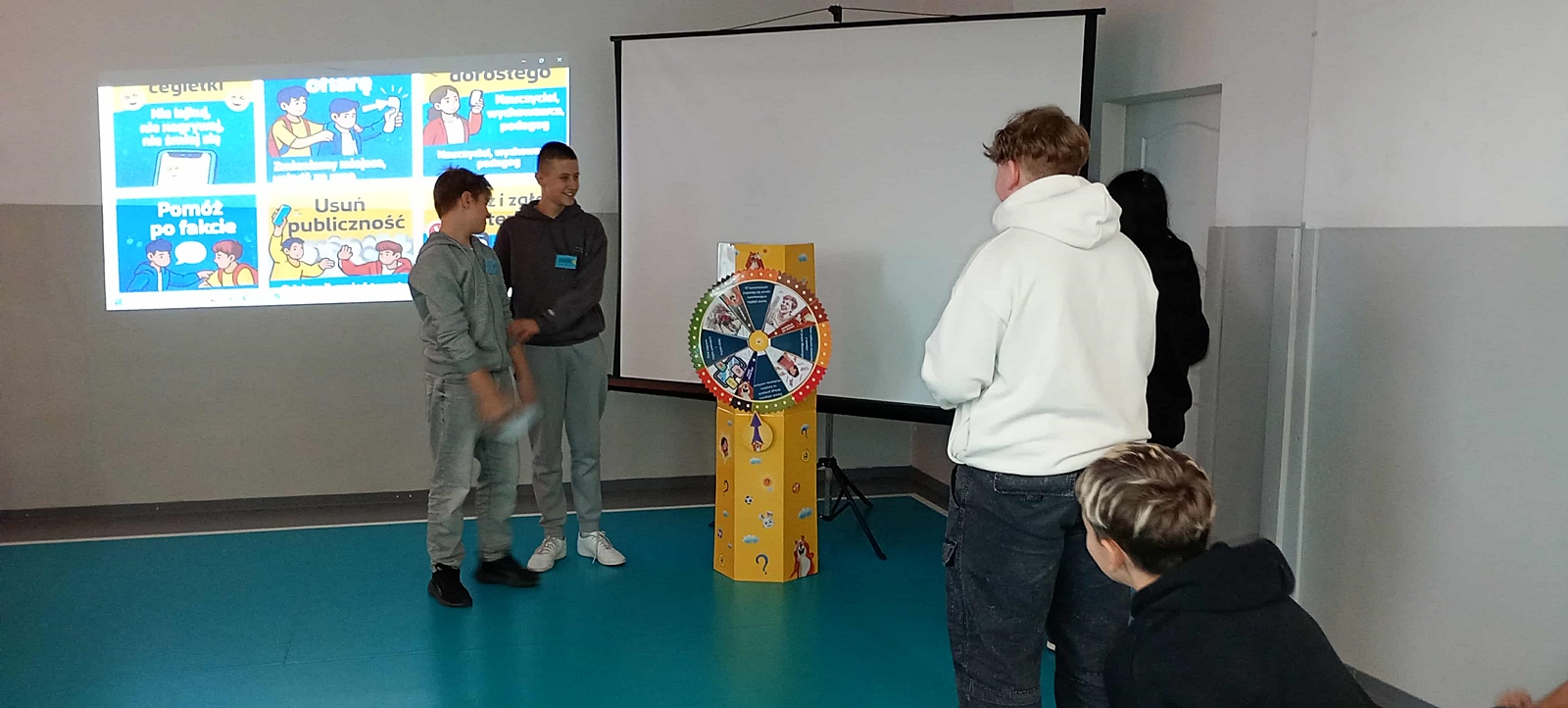 Grupa dzieci biorących udział w inscenizacji. W tle, na ekranie projekcyjnym wyświetlony jest slajd zawierający rady dotyczące zwalczania hejtu: Pomóż po fakcie, Usuń publiczność.
