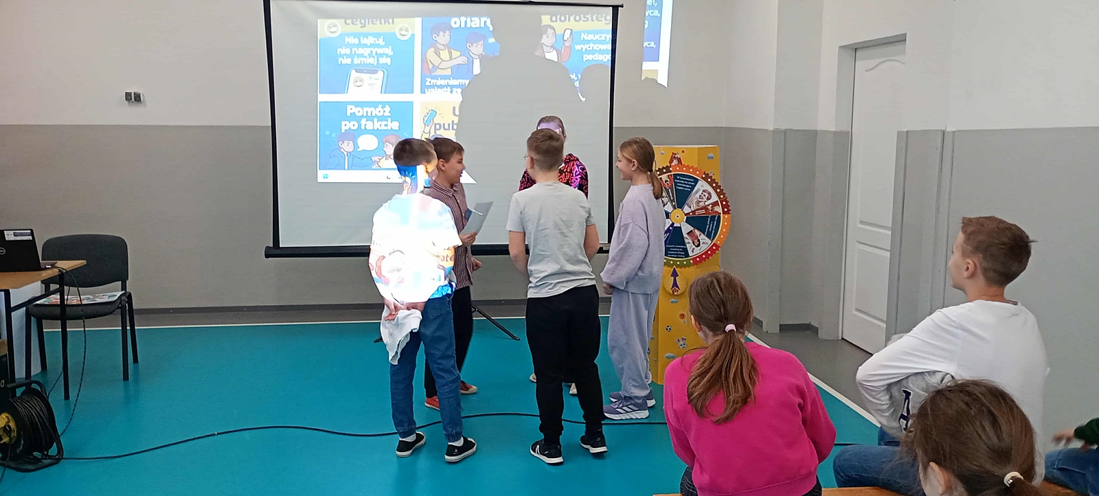 Grupa dzieci biorących udział w inscenizacji. W tle, na ekranie projekcyjnym wyświetlony jest slajd zawierający rady dotyczące zwalczania hejtu: Pomóż po fakcie, Usuń publiczność.