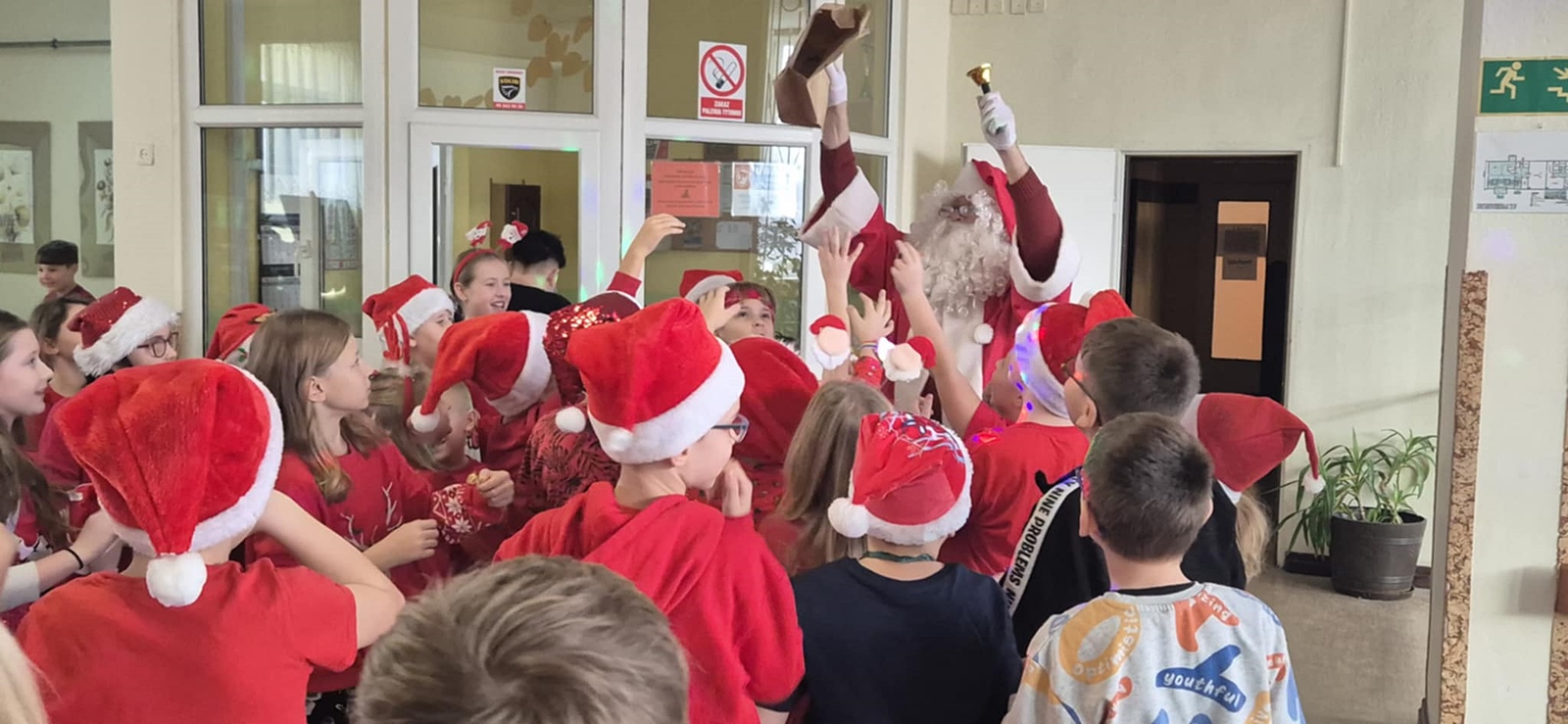 Grupa dzieci w czerwonych czapkach Mikołaja, zebranych wokół osoby przebranej za Świętego Mikołaja. Zdjęcie zrobiono w szkole.