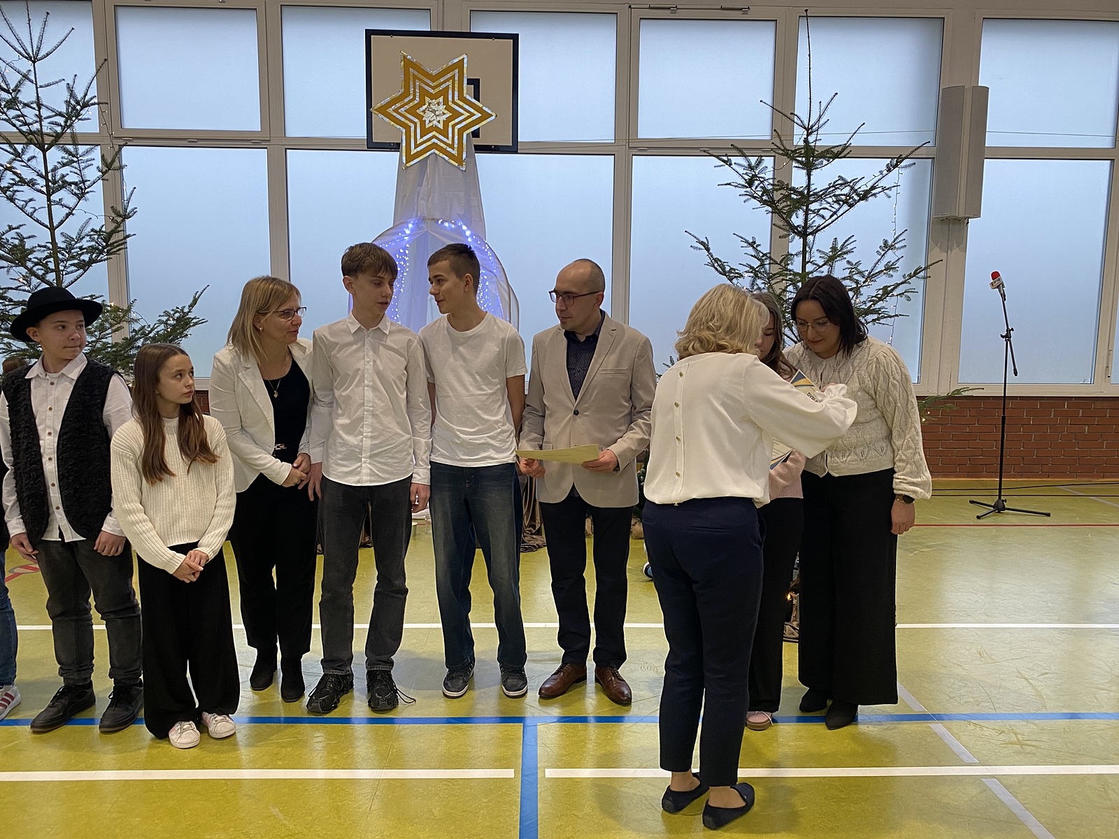 Grupa odświętnie ubranych osób zgromadzonych na świątecznej uroczystości, na której wręczane są dyplomy za udział w konkursie. W tle żywe choinki i duża ozdoba w kształcie gwiazdy.