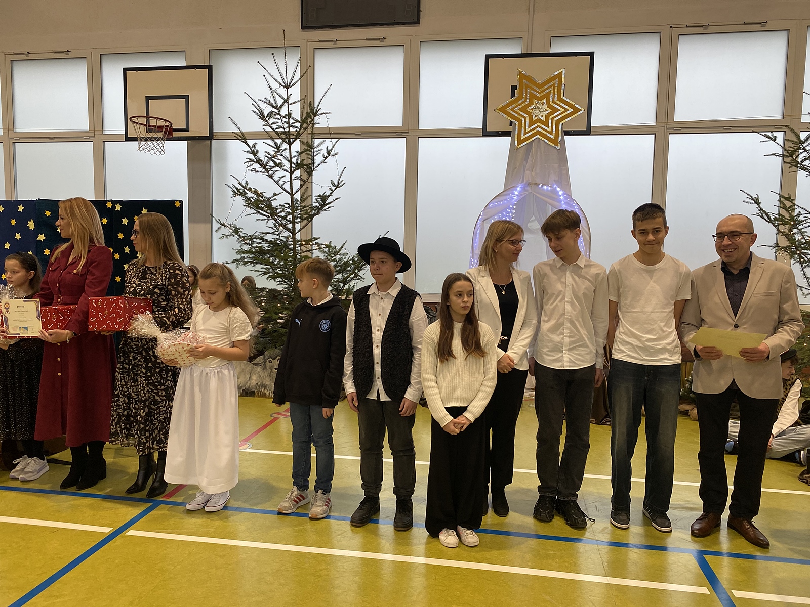 Na udekorowanej świątecznie scenie stoi grupa dzieci i dorosłych. Trzymają w rękach dyplomy i małe prezenty.