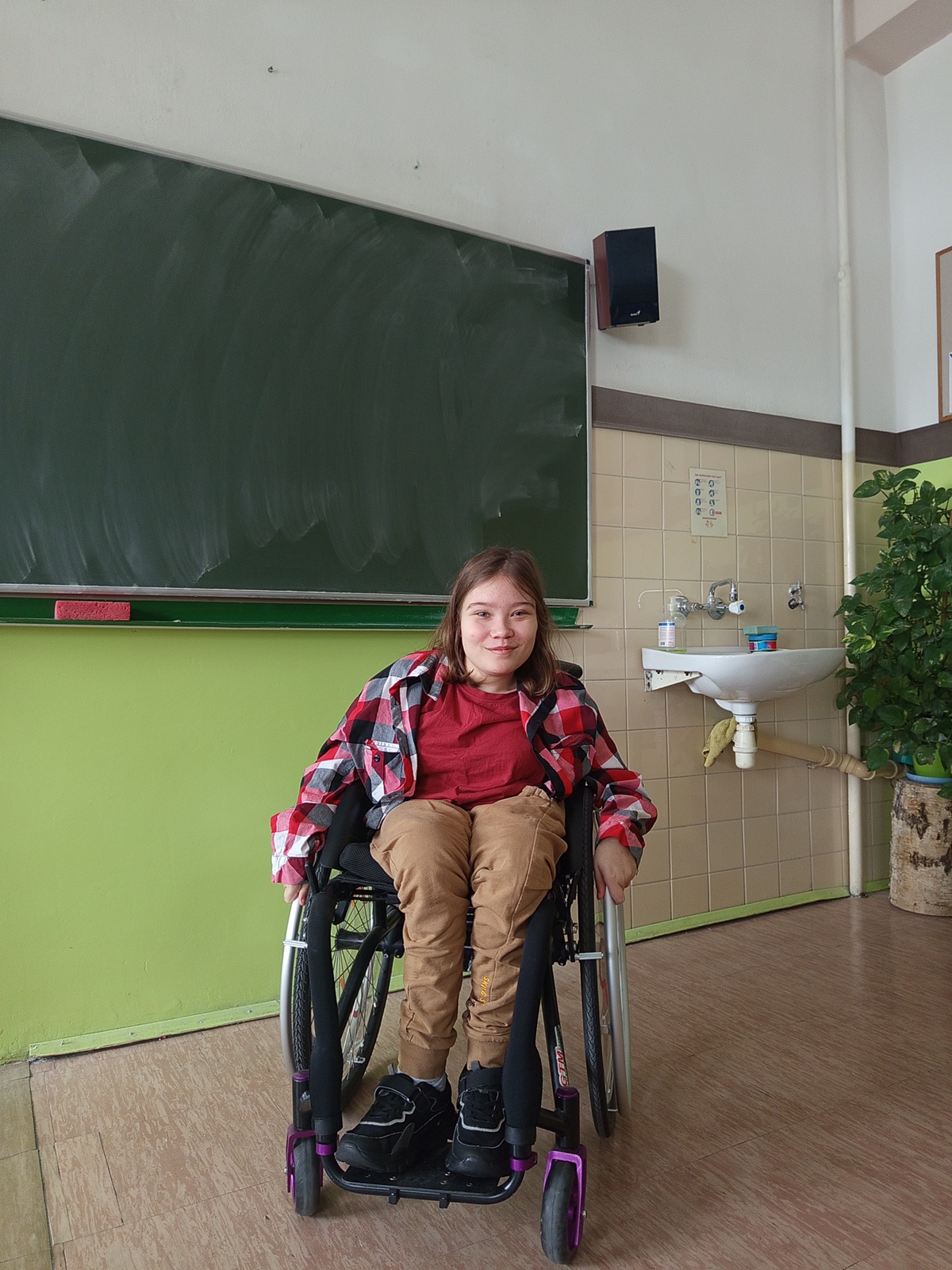 Uśmiechnięta uczennica na wózku inwalidzkim ubrana w kolorową koszulę.