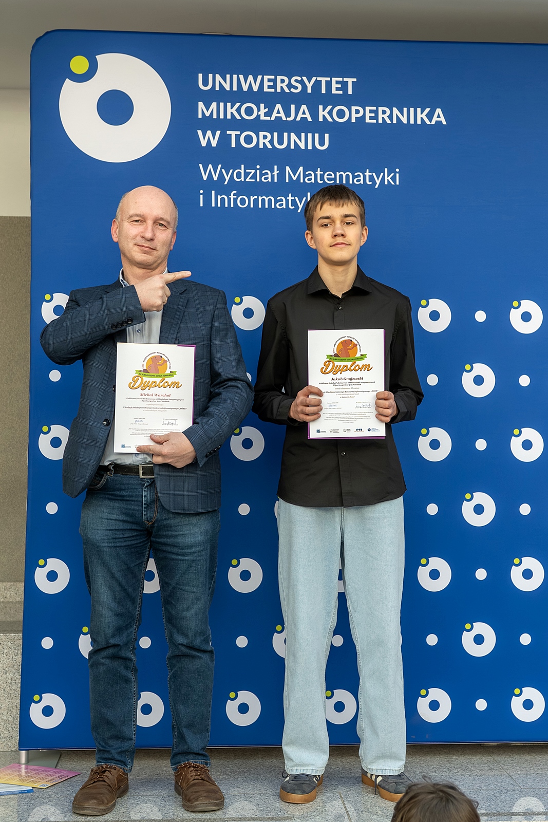 Nauczyciel z nagrodzonym uczniem. Nauczyciel wskazuje palcem na ucznia. W dłoniach dyplomy. Za ich plecami niebieskie tło z logo wydziału Matematyki i Informatyki.