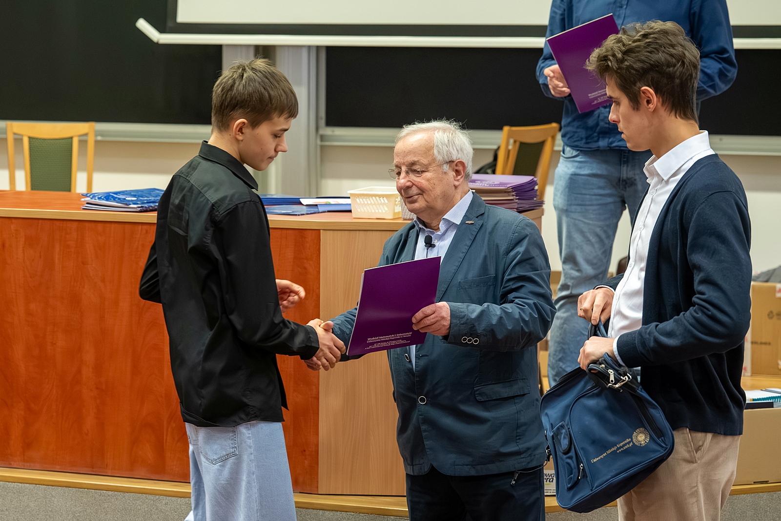 Moment wręczania dyplomu. Uścisk dłoni. Profesor wręcza dyplom nagrodzonemu uczniowi. Asystent profesora trzyma w dłoni nagrodę.