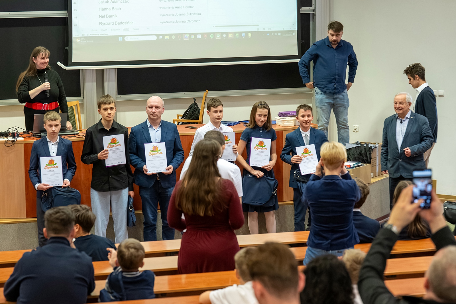Zdjęcie grupowe nagrodzonych uczestników konkursu. Sześć osób w rzędzie, trzyma w rękach dyplomy. W tle katedra wykładowa i fragment ekranu.