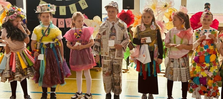 Grupa dzieci stojących na scenie podczas obchodów "Pierwszego Dnia Wiosny". Dzieci ubrane są w ekologiczne stroje wykonane z materiałów z recyklingu.