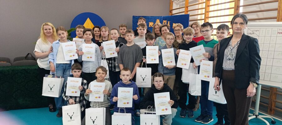 Grupowe zdjęcie uczniów i dorosłych w sali gimnastycznej. Wszystkie dzieci trzymają w rękach dyplomy i torebki upominkowe.
