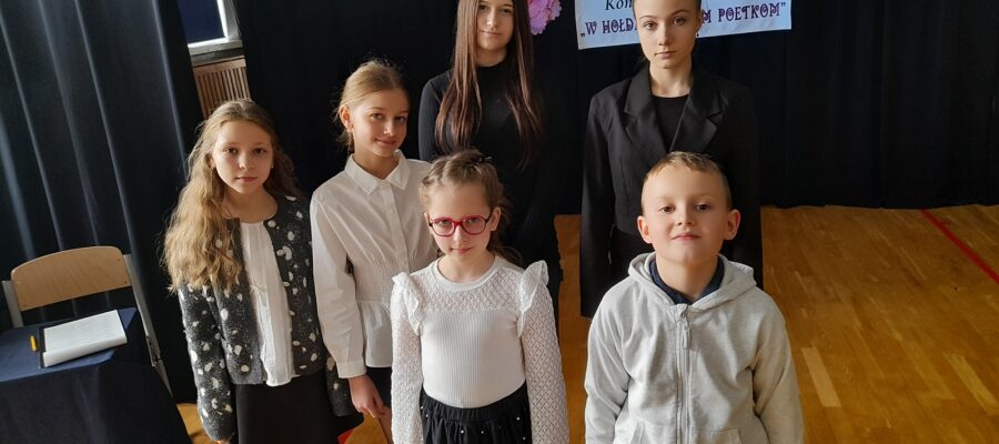 Grupa sześciu osób - pięcioro dzieci i nastolatka, pozują w sali gimnastycznej. W tle napis "Konkurs recytatorski: W hołdzie polskim poetkom".