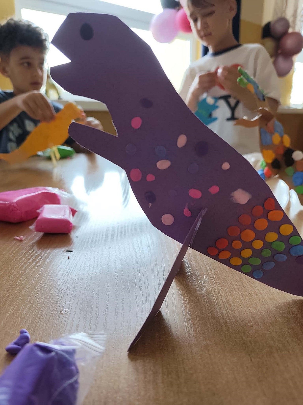 Wycięty z papieru fioletowy dinozaur, ozdobiony przez dzieci kolorowymi kropkami z piankoliny. W tle dwie postaci dzieci zdobiące swojego dinozaura piaknoliną.